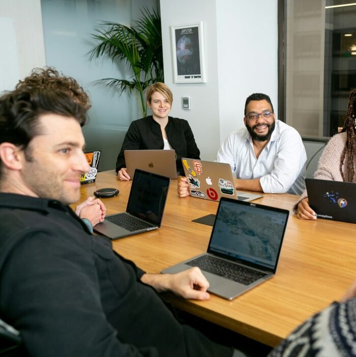 four people all on laptops, two men and two women, listen to person talking in a board meeting